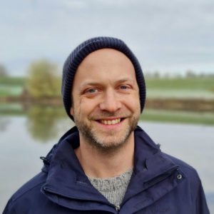 A smiling man wearing a dark beanie and navy jacket stands outdoors by a calm body of water, with green fields and trees blurred in the background under a cloudy sky.