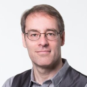 A man with short brown hair and glasses, wearing a grey collared shirt and a dark waistcoat, poses against a plain white background.