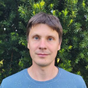 A man with short brown hair and blue eyes stands in front of dense green foliage, wearing a light blue shirt and looking directly at the camera with a neutral expression.
