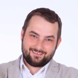 A man with short brown hair and a beard is smiling at the camera. He is wearing a light-coloured blazer over a white shirt and is posed against a plain white background.
