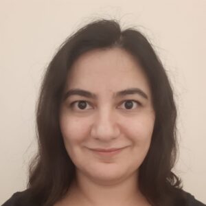 A person with long dark hair and dark eyes smiles softly at the camera. They are in front of a plain light-coloured background and wearing a dark top.