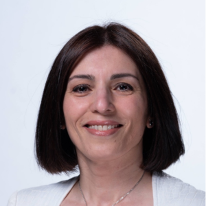 A woman with straight, dark brown shoulder-length hair smiles at the camera. She is wearing a white top and a necklace, and the background is plain white.
