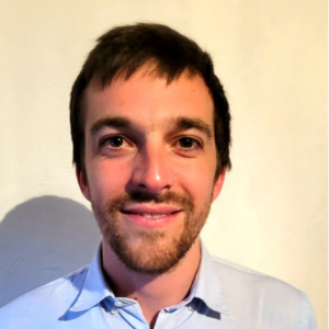 A man with short brown hair and a trimmed beard is smiling at the camera. He is wearing a light blue collared shirt and is standing against a plain, light-coloured background.