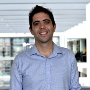 A man with short dark hair, wearing a light blue checked button-up shirt, smiles while standing indoors with modern architecture and large windows in the background.