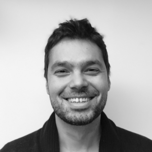 A man with short dark hair and a beard smiles at the camera against a plain white background.