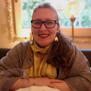 A woman with long brown hair, glasses, and large earrings smiles at a restaurant table. She wears a yellow scarf and grey jacket, with her hands resting on the table. A wine glass is in the foreground.