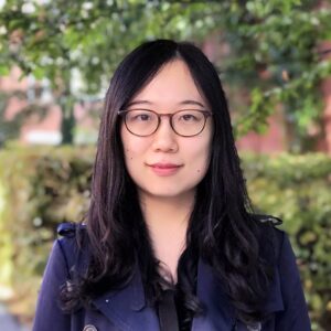 A woman with long black hair and glasses is standing outdoors, wearing a dark jacket. Green bushes and trees are blurred in the background.
