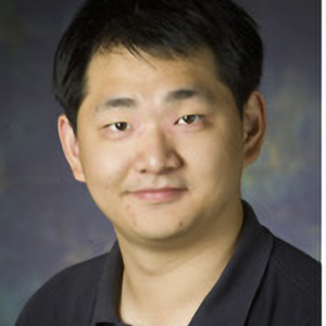 A man with short black hair and a slight smile is wearing a dark collared shirt. The background is softly blurred in shades of blue and green.