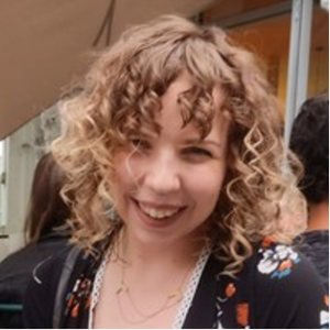 A young woman with curly blonde hair smiles at the camera. She is wearing a black floral-patterned top and layered necklaces, with people and a building blurred in the background.