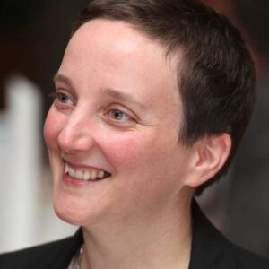 A person with short dark hair smiles whilst looking slightly to the side. They are wearing a dark jacket and the background is softly blurred.