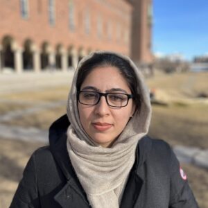 A woman wearing glasses and a beige headscarf stands outdoors on a sunny day, with a large brick building featuring arches in the background. She is dressed warmly in a black coat.