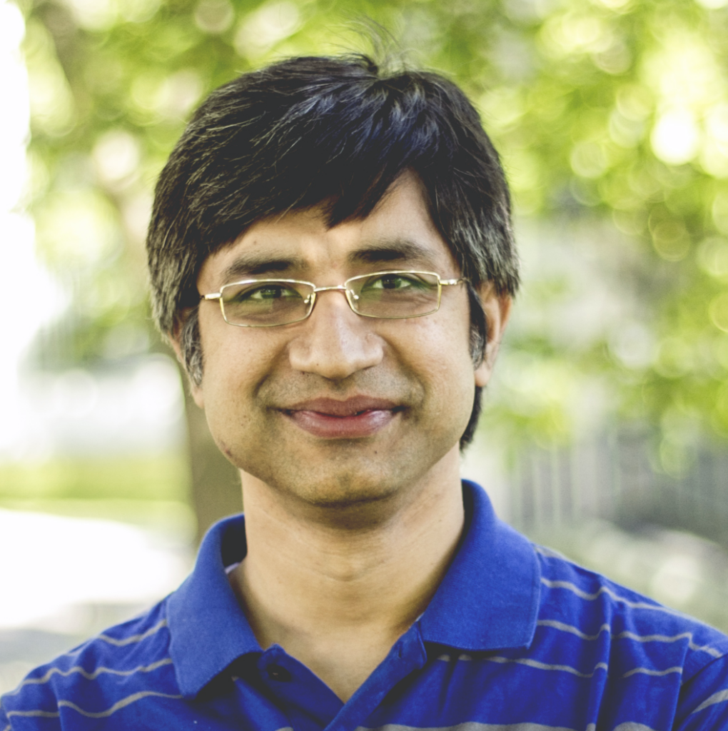A man with short dark hair and glasses smiles at the camera, wearing a blue striped polo shirt, standing outdoors with a blurred, green background.