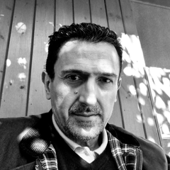 Black and white photo of a man with short dark hair and a trimmed beard, wearing a checked scarf and dark jacket, sitting in front of a wooden wall with dappled sunlight and shadow patterns.
