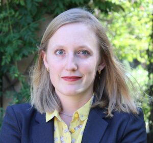 A woman with light blonde hair, wearing a dark blazer over a yellow shirt with a floral pattern, stands outdoors in front of greenery, looking directly at the camera with a neutral expression.