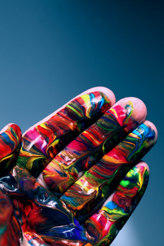 A close-up of a hand covered in bright, multicoloured paint swirls, with the fingers spread slightly apart against a smooth blue background.