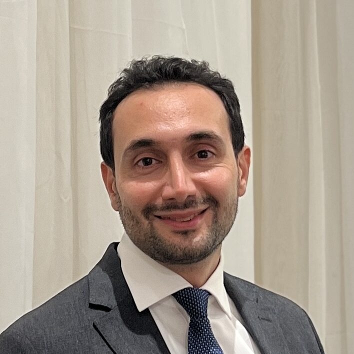 A man in a dark suit, white shirt, and blue tie with a pocket square stands in front of cream-coloured curtains, smiling at the camera.