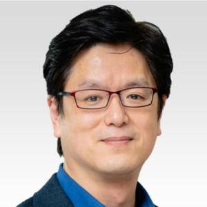 A man with short black hair and glasses, wearing a blue collared shirt and a dark blazer, smiles slightly against a plain light grey background.