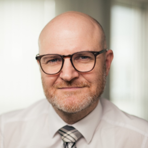 A middle-aged man with a bald head, light beard, and glasses is smiling slightly. He is wearing a white shirt, striped tie, and is in a softly lit indoor setting with blurred windows in the background.