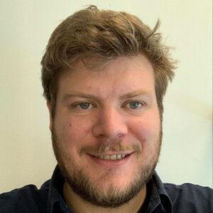 A man with light skin, short messy brown hair, and a beard smiles at the camera against a plain off-white background. He is wearing a dark collared shirt.
