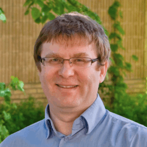 A man with light brown hair and glasses, wearing a light blue collared shirt, smiles whilst standing outdoors in front of green foliage and a tan brick wall.