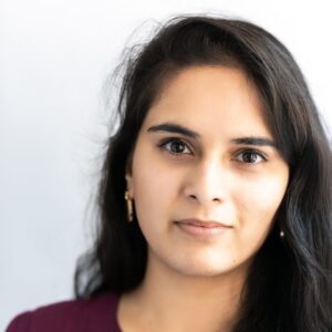 A woman with long dark hair and brown eyes looks directly at the camera with a neutral expression, wearing a purple top and gold earrings, set against a light, blurred background.