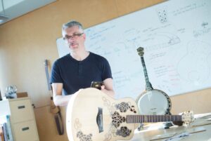 A man with grey hair and glasses stands in a bright room, holding a mobile, with a guitar and banjo on a table in front of him. A whiteboard filled with musical instrument sketches and notes is in the background.