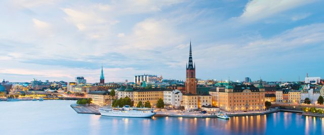 Panoramic view of Stockholm’s waterfront at dusk, with historic buildings, a prominent church spire, and boats moored along the water under a partly cloudy sky.