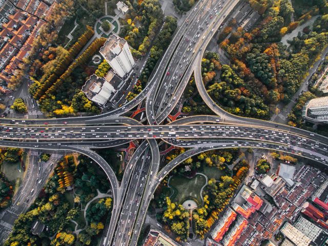 Aerial view of a large, busy multi-level motorway interchange surrounded by trees, buildings, and city streets, with cars densely travelling in all directions.