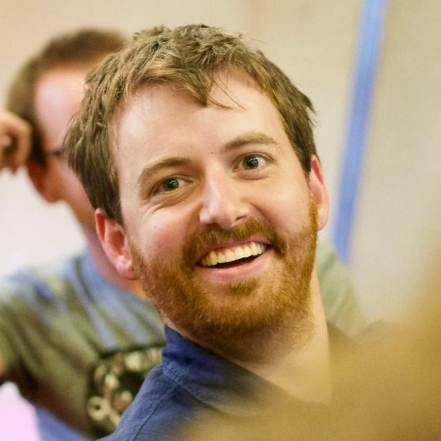 A man with a reddish-brown beard and short hair is smiling widely, looking slightly off camera. He is wearing a blue shirt, and another person is blurred in the background. The overall mood is cheerful and lively.