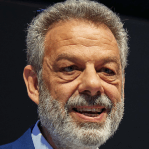 A middle-aged man with grey hair and a full beard is smiling. He is wearing a blue suit jacket and a light shirt. The background is dark and out of focus.