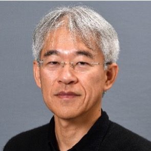A middle-aged man with short grey hair and glasses, wearing a black shirt, is facing the camera and smiling slightly against a plain grey background.
