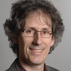 A man with wavy brown hair wearing glasses and a dark jacket smiles at the camera against a neutral background.