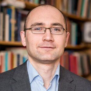 A man with short hair and glasses, wearing a grey suit jacket and striped shirt, stands in front of shelves filled with books. He is looking at the camera and smiling slightly.