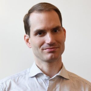A man with short brown hair, wearing a light-coloured, checked shirt, stands in front of a plain white background, looking at the camera with a slight smile.