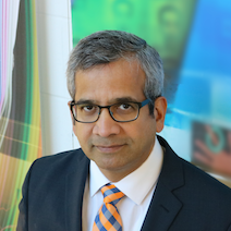 A man with short grey hair and glasses, wearing a dark suit, white shirt, and orange-and-blue striped tie, stands in front of a colourful, abstract background.