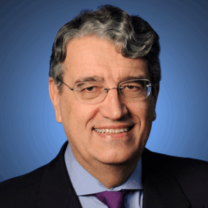 A smiling middle-aged man with grey wavy hair and glasses, wearing a dark suit, light blue shirt, and purple tie, posed in front of a blue background.