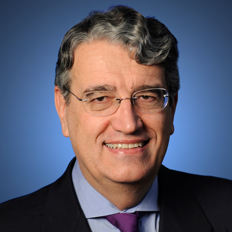 A smiling middle-aged man with grey wavy hair and glasses, wearing a dark suit, light blue shirt, and purple tie, posed in front of a blue background.