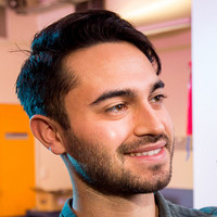 A young man with short, dark hair and a trimmed beard is smiling and looking slightly to the side in a brightly lit indoor setting.