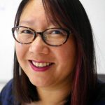 A woman with straight dark hair and glasses smiles at the camera. She is wearing bright lipstick and appears to be in an indoor setting with a light, blurred background.