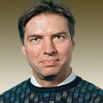 A man with short brown hair is wearing a patterned jumper over a collared shirt. He is facing the camera and smiling slightly, with a neutral, gradient background behind him.