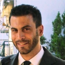 A man with short dark hair and a trimmed beard, wearing a suit and tie, is smiling outdoors with trees and a building in the background.