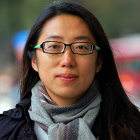 A woman with long dark hair and glasses, wearing a scarf and dark jacket, stands outdoors with a blurred background.