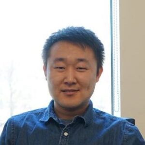 A person with short black hair wearing a denim shirt is sitting indoors in front of a bright window, smiling slightly at the camera.