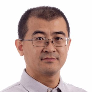 A man with short black hair, glasses, and a trimmed moustache and beard is wearing a light grey collared shirt and poses against a plain white background.