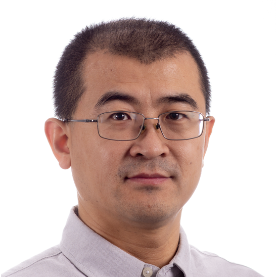 A man with short black hair, glasses, and a trimmed moustache and beard is wearing a light grey collared shirt and poses against a plain white background.