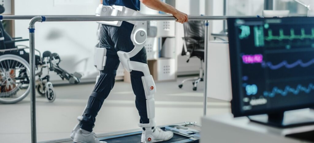 A person wearing a robotic exoskeleton walks on a treadmill whilst holding parallel bars in a rehabilitation facility; medical equipment and monitors are visible in the background.