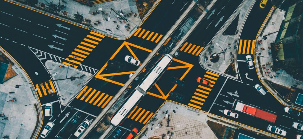 Aerial view of a busy city junction with multiple lanes, crossings, cars, buses, and yellow road markings. Pedestrians are visible on surrounding pavements. The scene is vibrant and dynamic.