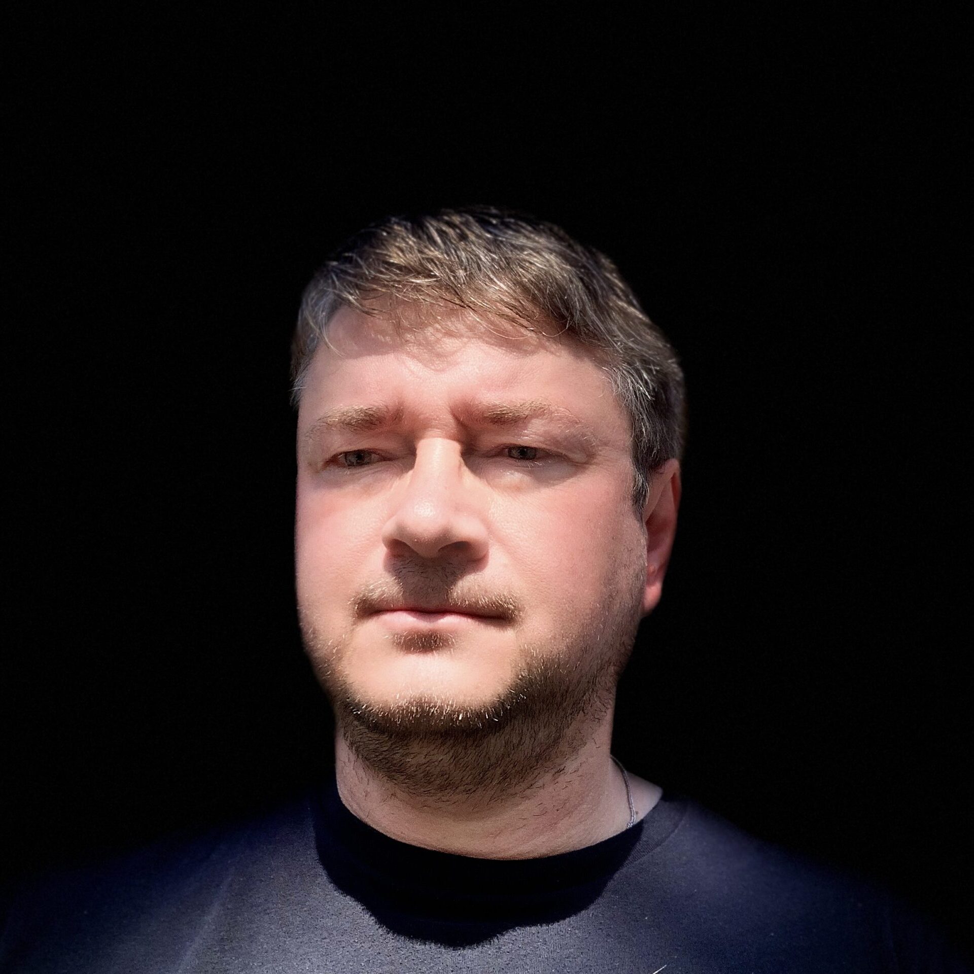 A man with short brown hair and a trimmed beard stands against a completely black background, wearing a dark shirt. Bright light shines on his face, casting sharp shadows.