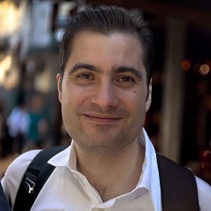 A man with short dark hair smiles at the camera. He is wearing a white collared shirt and has a black rucksack strap over his shoulder. The background is softly blurred, suggesting an outdoor urban setting.