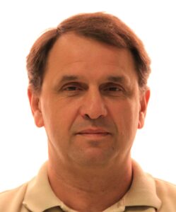 A middle-aged man with short brown hair wearing a light-coloured collared shirt is facing the camera with a neutral expression against a plain white background.